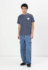 Camiseta de algodón gris con un logo de diamante menta en el frente, combinada con pantalones cargo de mezclilla azules holgados que tienen bolsillos laterales y zapatos negros.
