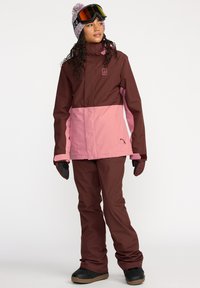 Conjunto de esquí que incluye una chaqueta marrón y rosa con un alto cuello, combinada con pantalones marrones. Los accesorios incluyen guantes negros y gafas de esquí.