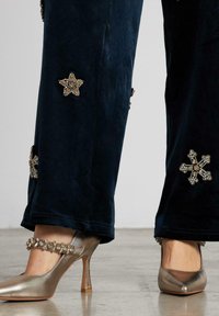 Los pantalones de terciopelo en azul marino oscuro presentan adornos decorativos en forma de estrellas y cruces doradas. Los zapatos de tacón alto metálicos dorados con tiras en el tobillo complementan el look.
