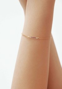 Pulsera de oro con una barra horizontal incrustada de piedras preciosas moradas, montada sobre una fina cadena, descansando en una muñeca de tono de piel.