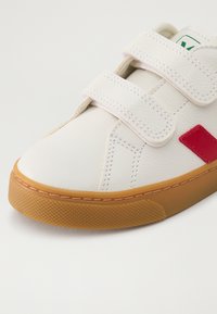 Baskets en cuir blanc avec deux sangles à boucle velcro, accent rouges et une semelle en caoutchouc marron présentant un motif texturé.