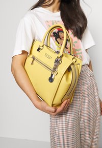 Nainen, joka pitää kädessään keltaista Guess-laukku, jossa on hopeiset yksityiskohdat, pukeutuneena valkoiseen graafiseen t-paitaan ja ruutuhousuihin, seisoo yksinkertaista taustaa vasten.