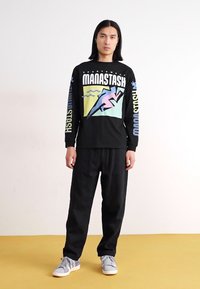 Black long-sleeve shirt with colorful graphic print, paired with black pants and gray sneakers. Background is white, and floor is yellow.