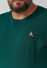 Behaarde man in een donkergroen T-shirt met ronde hals en een geborduurde vliegende eend op de linkerborst.