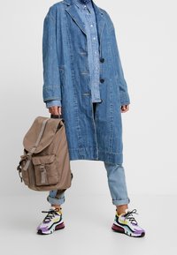 Personne portant une veste en jean superposée et un jean, tenant un sac à dos marron, avec des baskets colorées sur fond blanc.