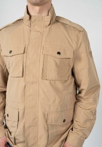 Giacca leggera beige con colletto alto, dotata di due tasche sul petto e due tasche inferiori, realizzata in materiale liscio con dettagli in bottone.