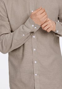 Camicia a maniche lunghe marrone chiaro con davanti a bottoni, bottoni bianchi e un tessuto morbido e texturizzato. I polsini sono regolabili con uno spacco.