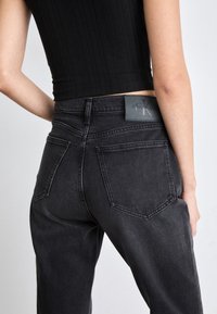 Black high-waisted jeans with a straight leg, featuring a Calvin Klein leather patch on the back waistband and double back pockets.