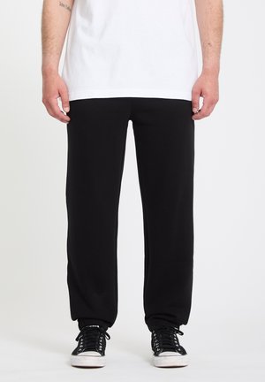 Personne portant un pantalon de jogging noir, des baskets noires et blanches, et un t-shirt blanc, debout devant un fond blanc uni.