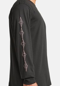 Langärmliges schwarzes Shirt aus Baumwolle mit pinkfarbener, diamantförmiger Stickerei entlang des linken Ärmels. Glatte Textur, lässiges Design.