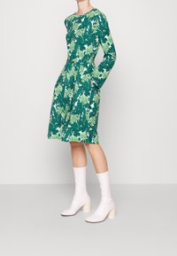 Abito verde a fiori con maniche lunghe, vita aderente, gonna svasata, tasche laterali, abbinato a stivali con tacco alto rosa pallido.