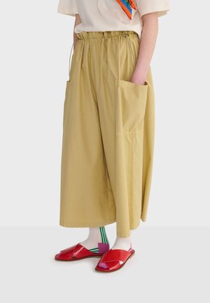 Szerokie beżowe spodnie z dużą boczną kieszenią, elastycznym pasem, noszone z białymi skarpetkami z zielonymi paskami i czerwonymi sandałami z otwartymi palcami.