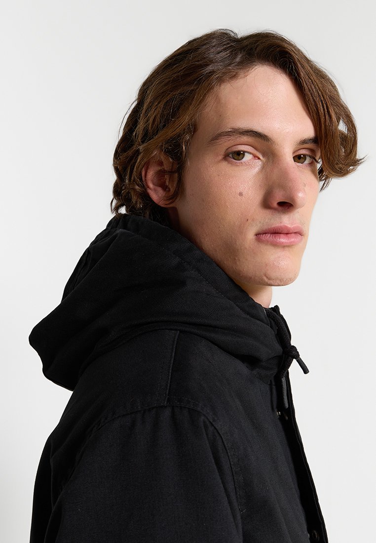 Veste à capuche noire avec cordon de serrage, présentant une texture lisse et une coupe décontractée. Le modèle regarde à gauche contre un fond neutre.