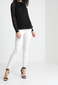 Top lungo a maniche lunghe plissettato nero con maniche trasparenti, abbinato a jeans skinny bianchi e tacchi neri open-toe. Completo semplice ed elegante.