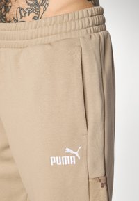 Tuvumā redzama bēšā Puma sporta bikses kabata un josta, ar redzamu tetovētu ādu virs biksēm.