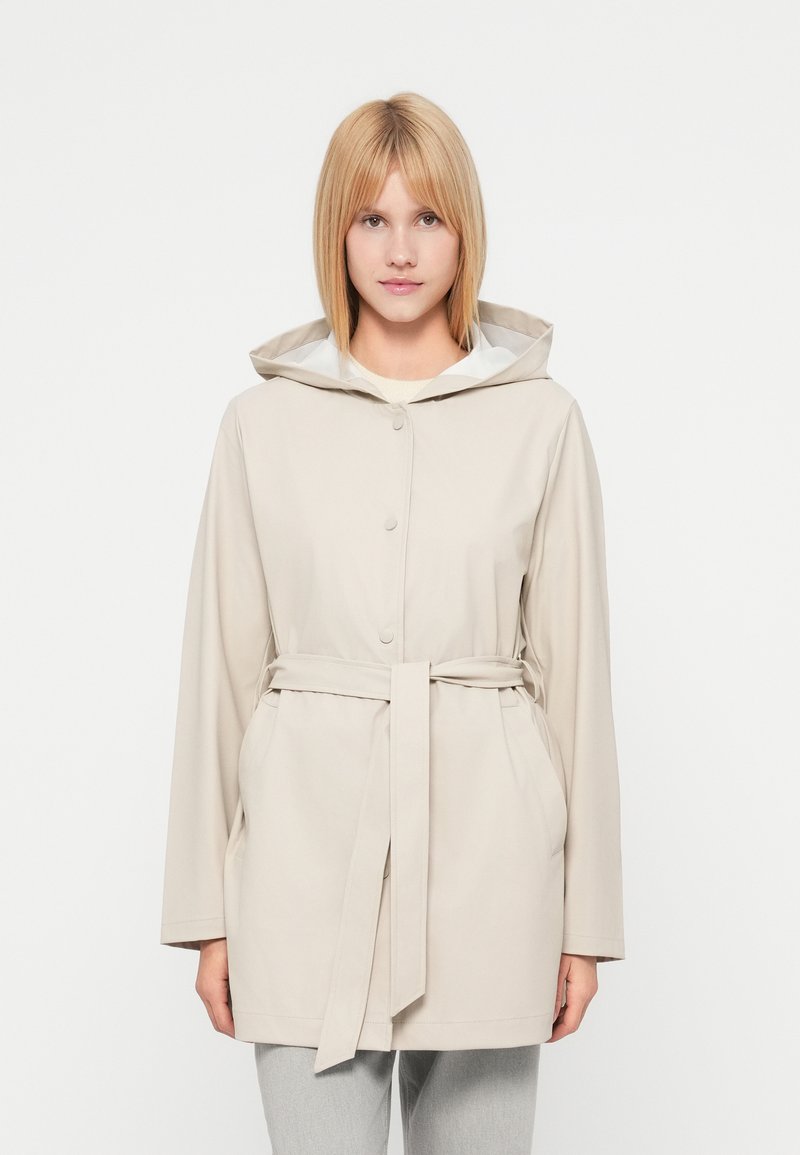 Veste imperméable beige avec capuche, fermeture avant à boutons, taille ceinturée et deux poches latérales. Texture douce, design décontracté.