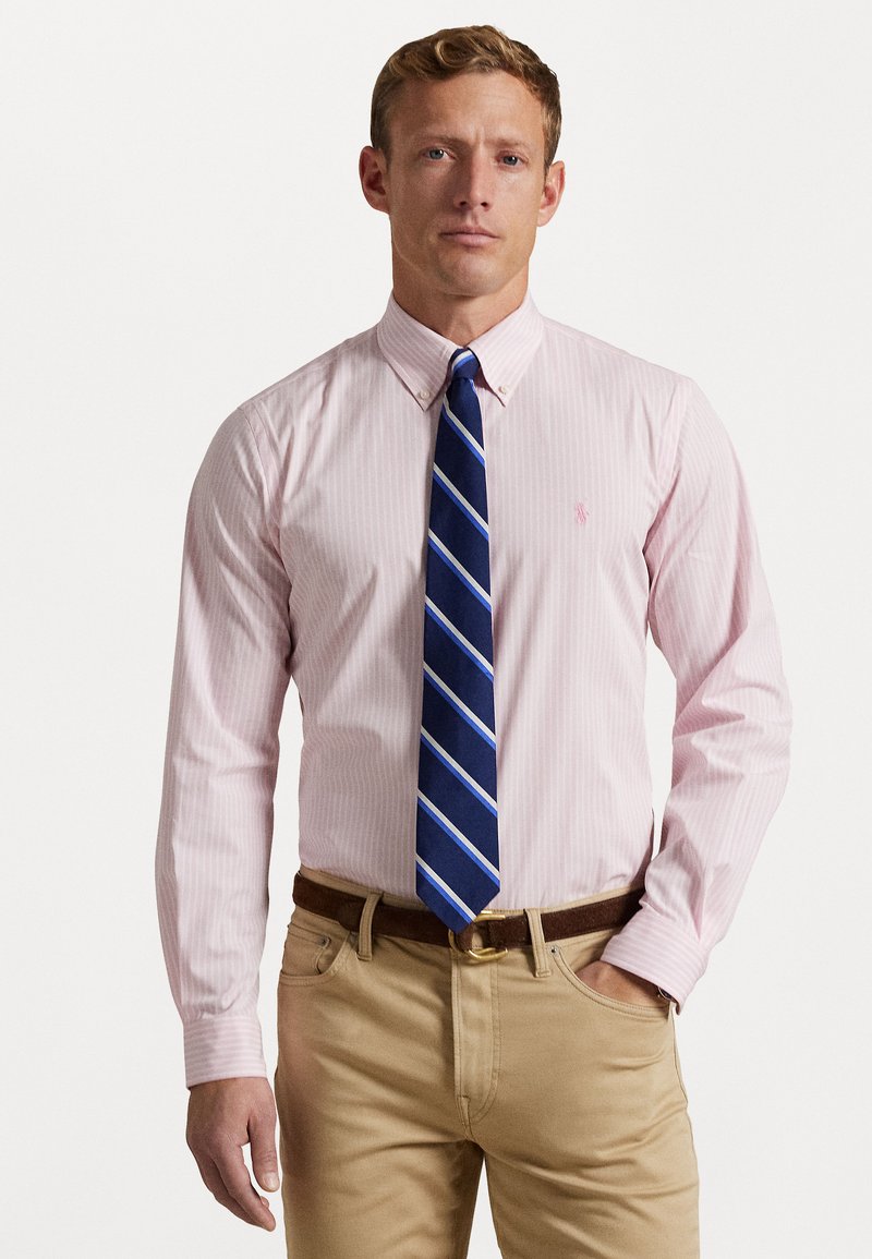 Camisa de botones a rayas rosas con cuello, combinada con una corbata a rayas azul y pantalones caqui. Presenta un cinturón marrón y un corte ajustado.