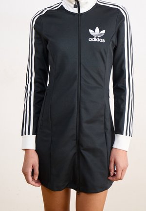 Robe zippée noire Adidas avec trois bandes blanches sur les manches, poignets blancs et logo Adidas sur la poitrine, portée par une personne.