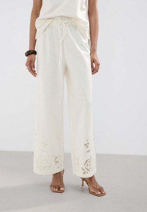 Mujer que lleva pantalones anchos color crema con patrones de recortes florales cerca del dobladillo, combinados con sandalias de tacón marrón claro y una blusa blanca.