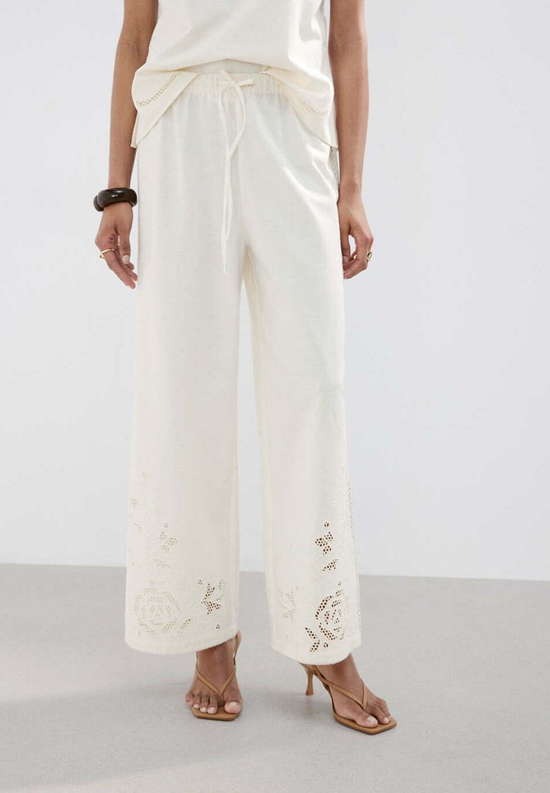 Femme portant un pantalon large crème avec des motifs floraux ajourés près de l'ourlet, associé à des sandales à talons couleur tan et un haut blanc.