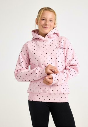 Jeune personne souriante, portant un sweat à capuche rose à pois et un pantalon noir, les mains doucement jointes devant sur un fond uni.