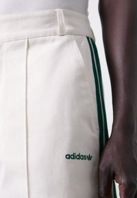 Vita manchesterbyxor med gröna tre ränder som detaljer. Har en broderad grön Adidas-logotyp på låret och en plisserad framdesign.