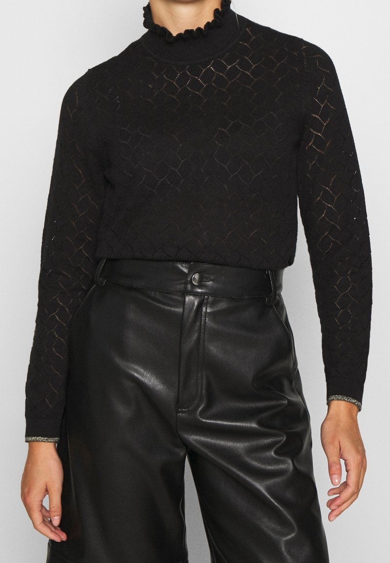 Femme portant un top en dentelle noire à manches longues avec un col festonné et un pantalon noir en simili cuir taille haute.