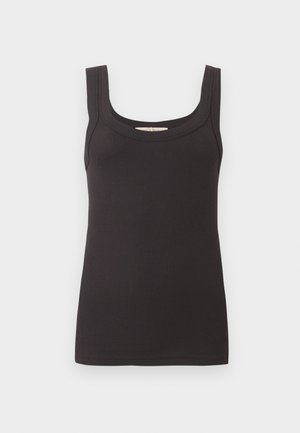 Femme portant un haut noir sans manches, un jean bleu à jambes larges et des sandales noires ouvertes à talons, debout devant un fond clair uni.