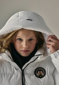 Doudoune blanche avec un col montant, fermeture éclair, et un patch logo rond sur le côté gauche. Elle possède une capuche et un tissu texturé.