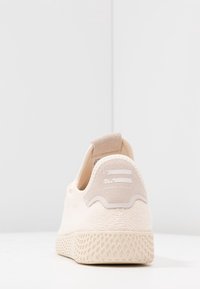 Ljusbeige sneaker med texturerad sula, mockahälflik och vita ränder ovanför texten "Pharrell Williams" på vit bakgrund.