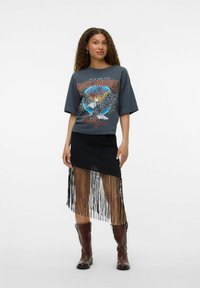 T-shirt gris graphique avec un design d'aigle et du texte, associé à une jupe noire avec des détails en fringes. Des bottes marron complètent la tenue.