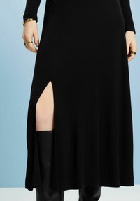Schwarzes Kleid mit einem seitlichen Schlitz bis zum Oberschenkel, aus glattem Stoff gefertigt. Verfügt über lange Ärmel und eine fließende Silhouette.