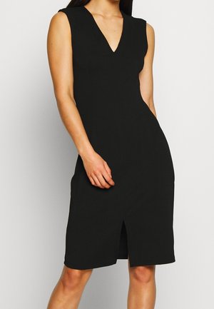 Femme portant une robe noire sans manches, à mi-genoux, avec un décolleté en V et une fente avant, se tenant devant un fond clair.