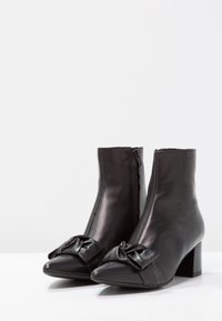 Botas de cuero negro a la altura del tobillo con tacones cuadradados y lazos decorativos en la punta, sobre un fondo blanco.