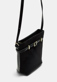Borsa a spalla in suede nera con tracolla in pelle sottile e regolabile, dettagli in metallo dorato e fibbia su sfondo bianco.