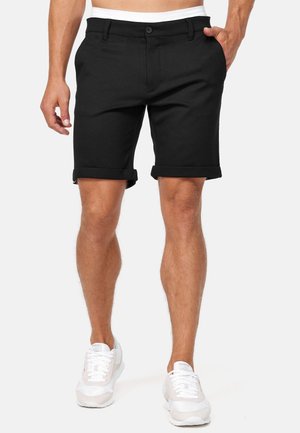 Homme portant un short noir avec ourlet retroussé et des baskets blanches, debout avec une main dans la poche devant un fond uni.