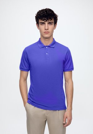 Jeune homme aux cheveux bouclés et foncés, portant un polo bleu à manches courtes et un pantalon beige, debout devant un fond blanc uni.