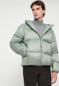 Jachetă puf verde deschis, cu glugă, quilt diagonal, fermoar frontal, buzunare laterale și un logo mic pe piept. Purtați peste un pulover de culoare gri.