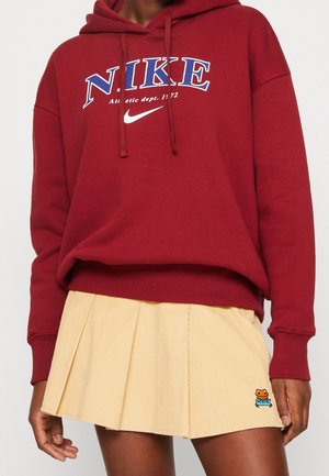 Personne portant un sweat à capuche rouge Nike avec un logo bleu et blanc et une jupe plissée beige avec un petit patch grenouille près de l'ourlet.