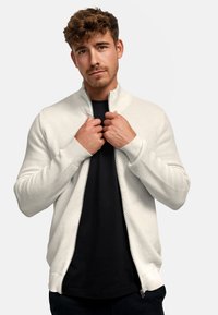 Jonge man met krullend haar die een witte ritsjack met structuur draagt over een zwart shirt, met beide handen de kraag van het jack vasthoudend.