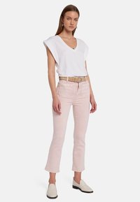 Haut blanc en V avec des manches bouffantes, jean rose clair coupe droite et ceinture beige. Le modèle porte des mocassins crème. Fond simple.
