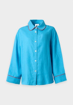 Femme portant une chemise ample bleu vif boutonnée et un pantalon large assorti avec un liseré rouge, debout sur un fond blanc uni.