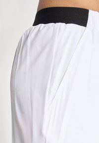 White athletic shorts with a black elastic waistband. The fabric is smooth and lightweight, featuring side pockets for utility.