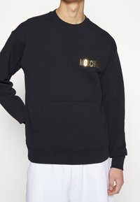 Černá mikina vyrobená z bavlny, má přední kapsu a zlatou fóliovou logo "MOSCHINO" na levé straně. Měkká textura, žebrované manžety.