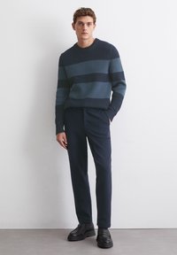 Gestreifter Pullover in Marineblau und Tannengrün mit gerippter Textur, kombiniert mit maßgeschneiderten Marineblauen Hosen und schwarzen Slippern.