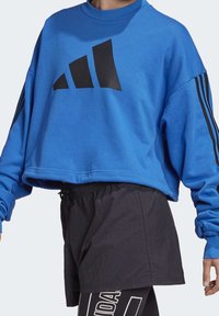 Sweatshirt bleu court en tissu doux avec un grand logo Adidas noir sur le devant, avec des bandes noires sur les manches. Associé à un short noir.