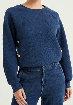 Kobieta ubrana w granatowy teksturowany sweter z długim rękawem z srebrnymi guzikami w połączeniu z ciemnoniebieskimi jeansami z wysokim stanem.