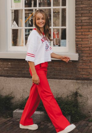 Jonge vrouw in rode broek en wit shirt loopt naast een bakstenen gebouw met een groot raam, glimlacht en kijkt naar de camera.