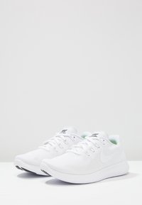 Zapatos deportivos blancos con parte superior de malla, punta redondeada y suela flexible. Incluyen cordones planos y un acento verde en el interior del talón.