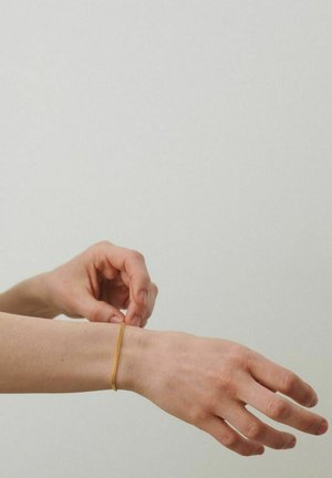 Roka piemeklē tievu zelta ķēdes aproci ap plaukstas locītavu uz gaiša vienveidīga fona.